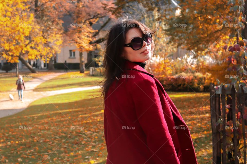 Portrait of a beautiful young caucasian brunette girl in sunglasses and a red demi-season coat stands half-turned in a colorful autumn public park, close-up side view. Concept autumn clothing demi-season.