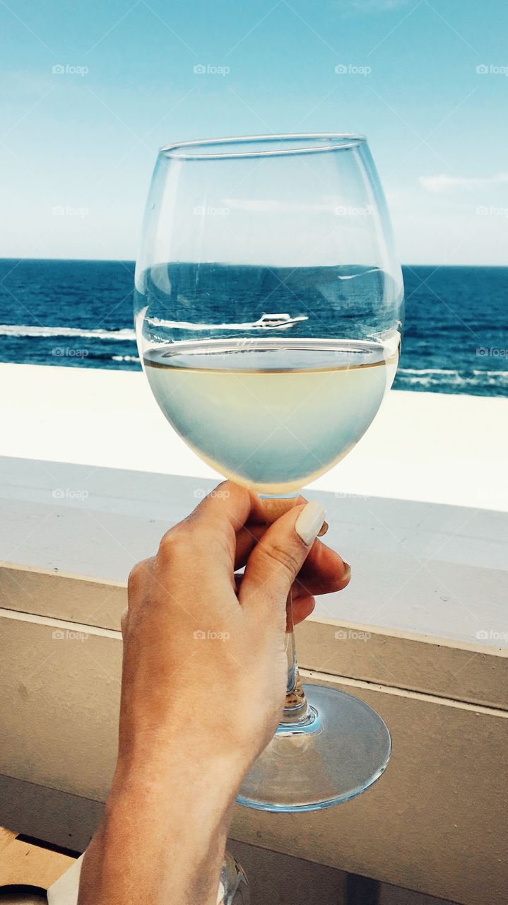 The hand holds a glass of white wine, against the background of the sea and a boat.