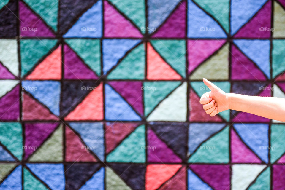 Person's hand giving a thumbs up into the frame of the image against a colorfully patterned wall