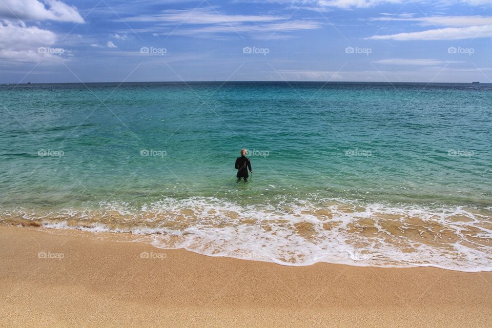 A lone girl in a wetsuit walking off a sandy beach and into an emerald green ocean.