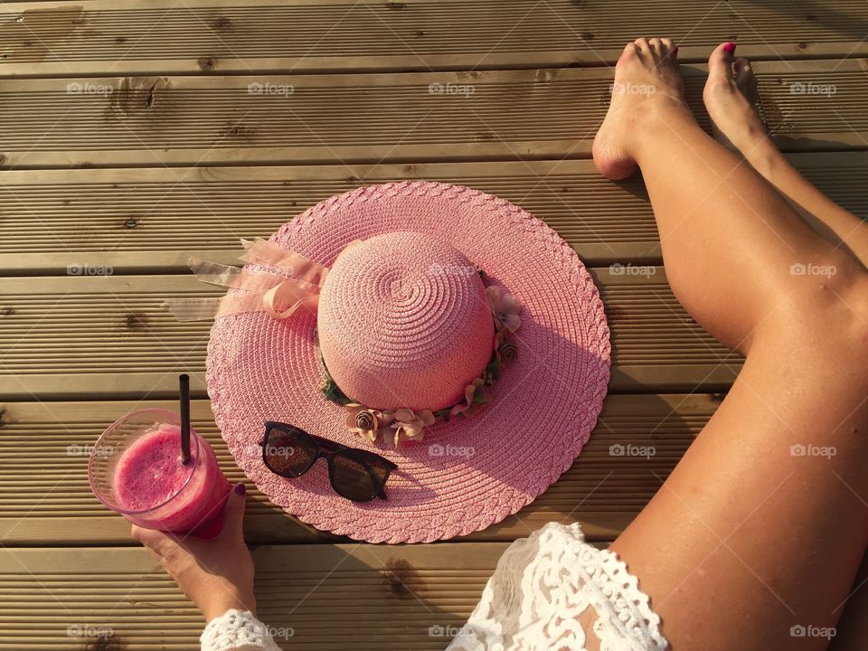 Woman's tanned sexy legs on wooden pontoon with glass of raspberry smoothie and pink summer hat 