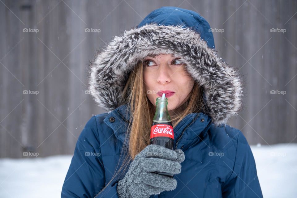 Smiling young woman wearing a winter coat and playfully drinking from a bottle of soda from a straw 