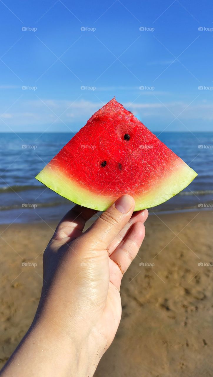 Hand holds a triangular shape watermelon slice on the background of the sea