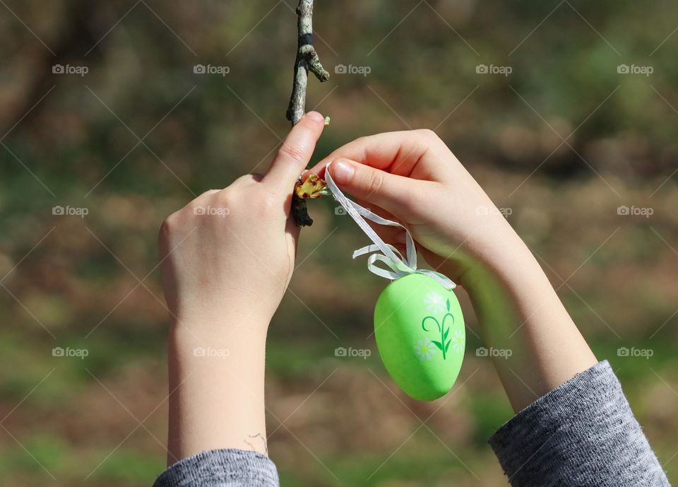 The hands of a little caucasian girl hang a decorative easter egg green with an ornament on a tree branch, side view close-up. Holiday tradition concept.