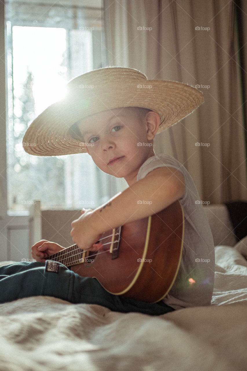 Toddler boy playing on ukulele guitar in straw hat