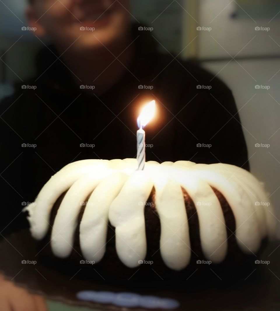 A young adult man celebrating his birthday with a smile and a bunt cake with cream cheese icing and one candle flame on top in a kitchen