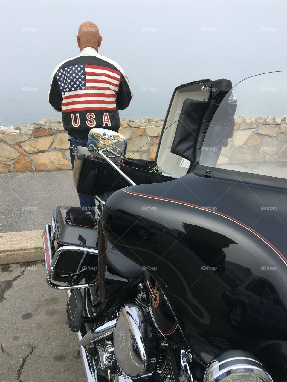 American Flag on back of leather jacket worn by motorcycle biker standing behind black Harley Davidson Dresser with trunk open.