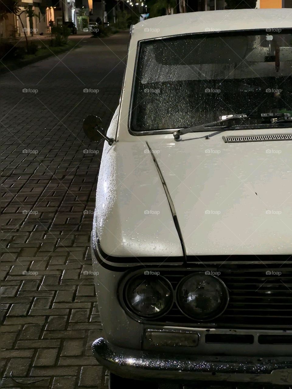Vintage white car parked on a dark street with some lights lighting it up, in the middle of the night.