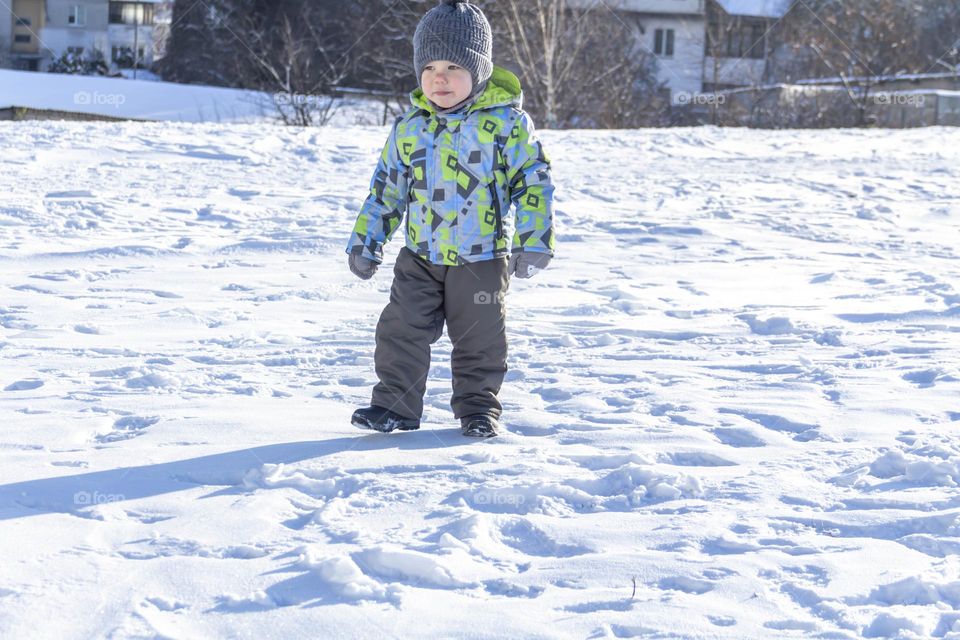 A child in winter in winter jackets, pants, a hat and boots on white snow on the street and in the park in nature plays winter fun and sleds.
