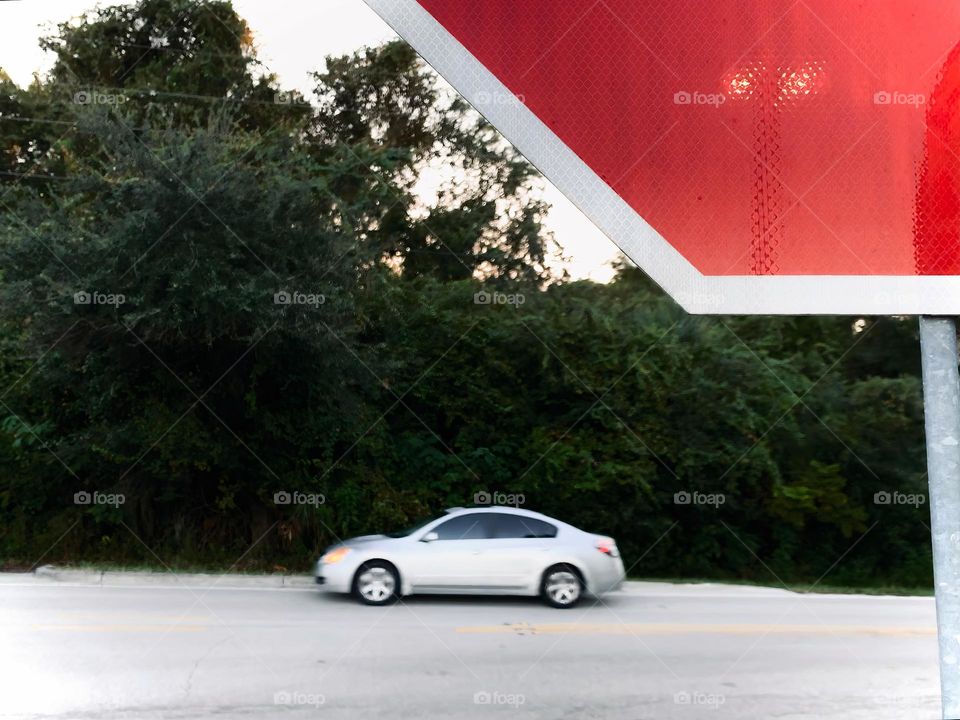 Commuting: cars driving by the a small mall and grocery store, seen from a stop sign with bushes and trees.