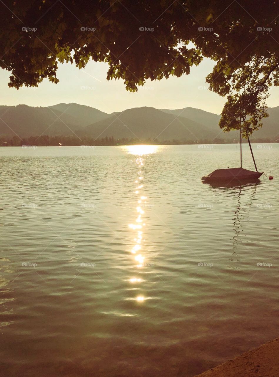 The sunset, sun shone down the water in a long yellow streak,an moored boat,many mountains and trees in front , I was standing on the edge of a lake with foliage  reach out the water.Very beautiful yellow light glittering on lake water surface
