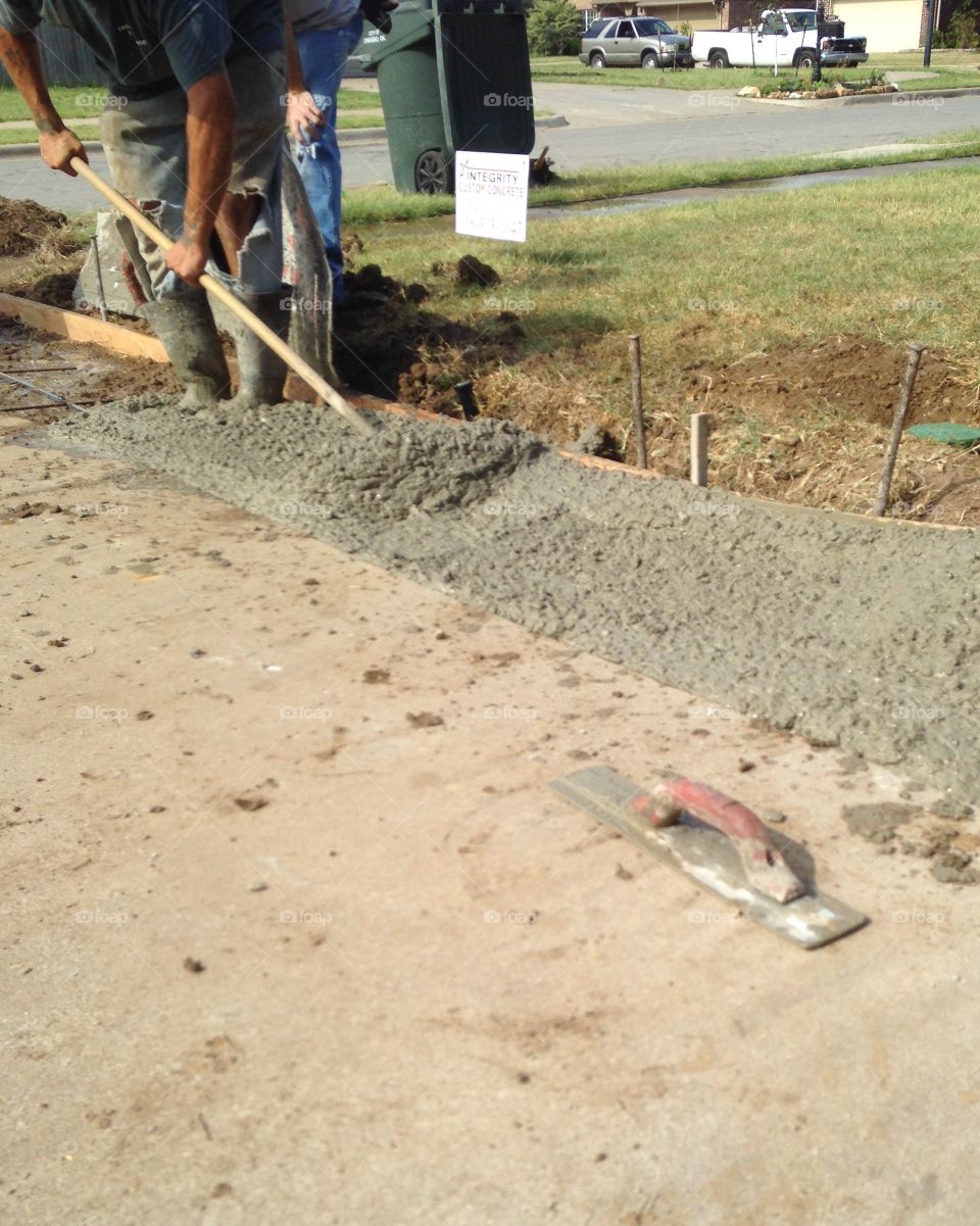 Construction of cement sidewalk shows a trowel in foreground and a man shoveling wet cement.