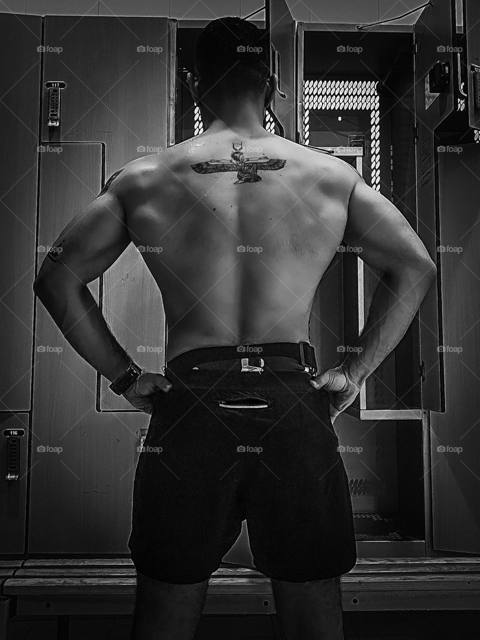 A man seen from the back at a gym locker room, with a tattoo of the Ancient Egyptian Goddess Eset (also known as Aset or Isis) on his back, with some other less visible tattoos on his left arm. 