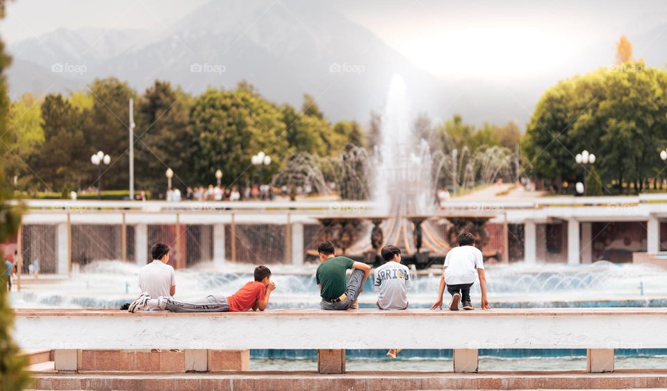 Boys relaxing close to a huge water fountain from park and admiring the mountain view