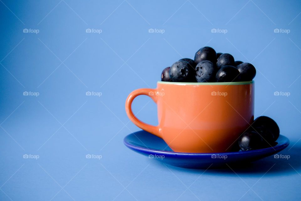 Orange Teacup filled with Blueberries on a Blue saucer with blue background
