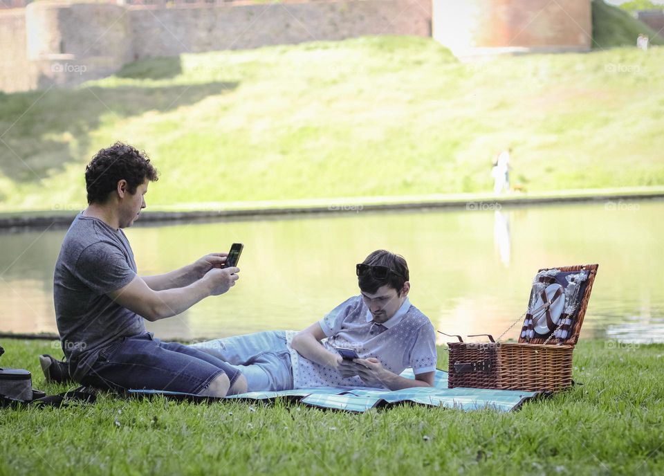 Two young caucasian men relaxing with mobile phones at a picnic near a lake in a city park on a summer day, side view close up. Male picnic concept.