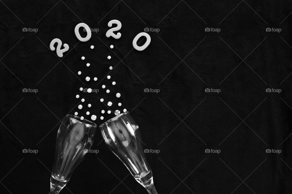 Flat lay of the numbers ‘2020’ above champagne glasses with rhinestone bubbles on black background in black and white - copy space on right side of landscape format