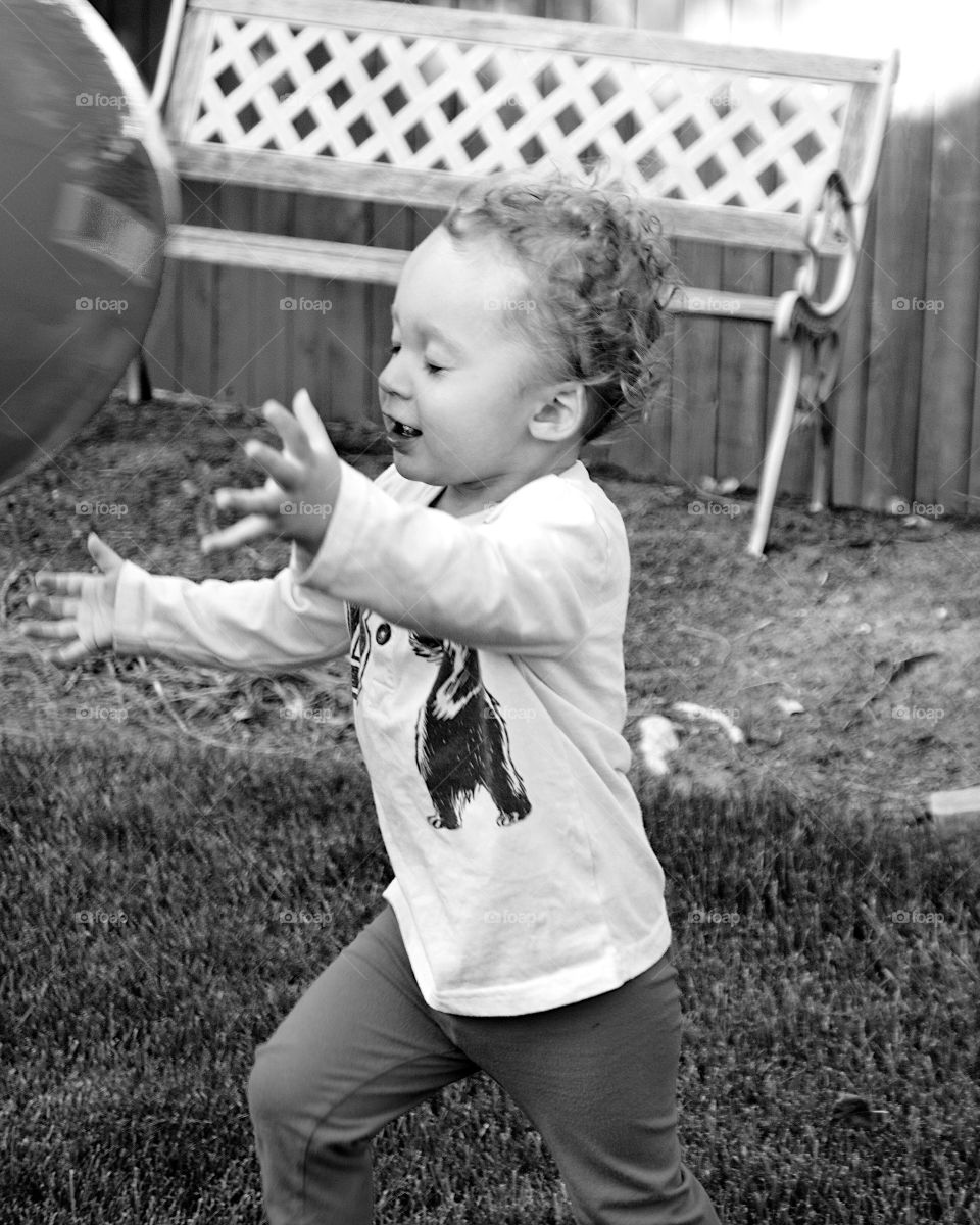 A small toddler boy in contrast to  a giant rubber ball he is trying to catch on a sunny summer day in the backyard. 