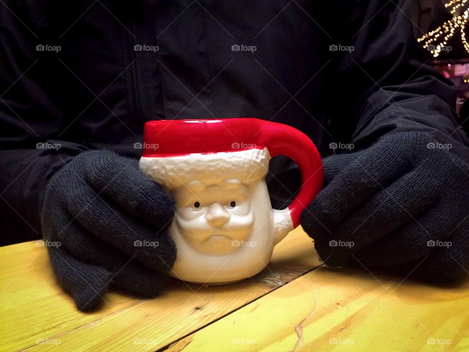 Man’s hands wearing black wool gloves holding a Santa Claus shaped mug with Christmas lights in the background 
