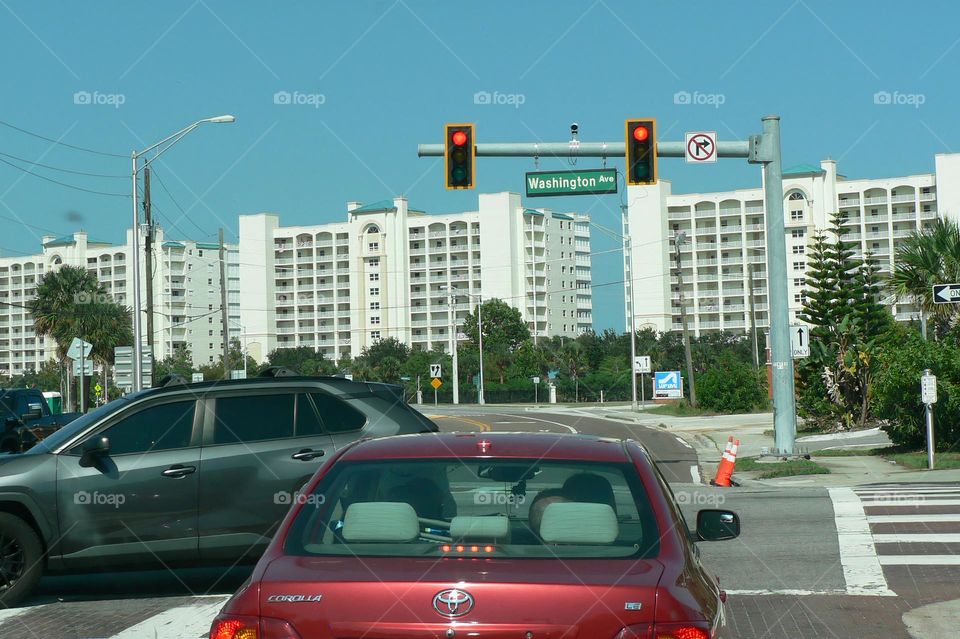 Commuting: traffic at an intersection with traffic lights with red car in front and grey car turning left, all in front of many white tan apartments buildings and trees.