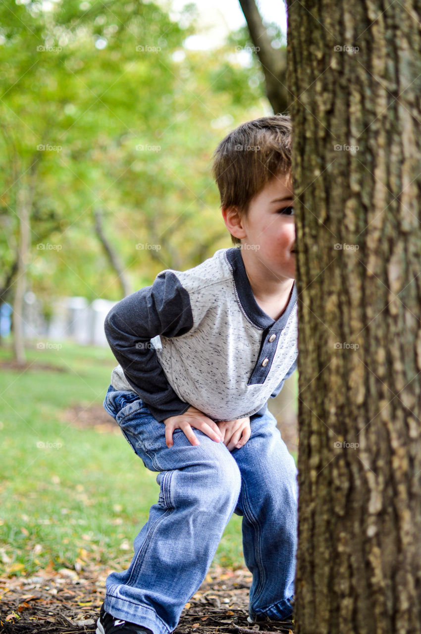 Toddler boy playing hide and seek behind a tree at the park