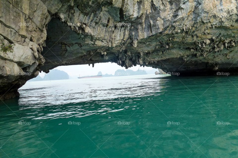 I am sailing on a bay,beautiful blue water color,In the foreground is a rocky cave,I will row there to explore.in the distance there is a large ship and some mountains.I won't be able to go out there by boat because the waves are very strong