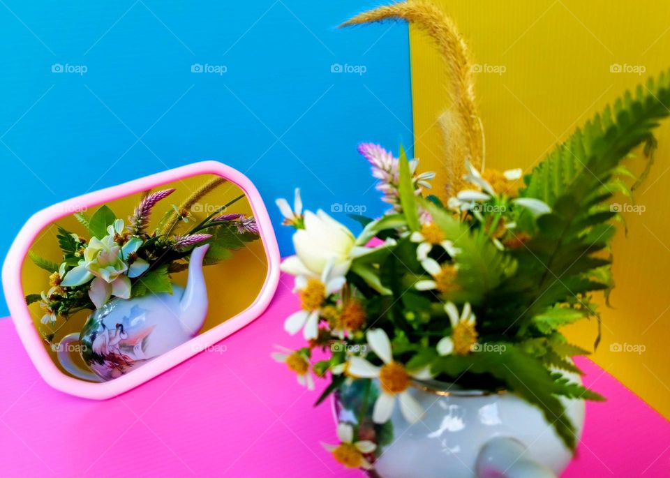 A teapot of flowers in the mirror on pink, yellow and blue background.