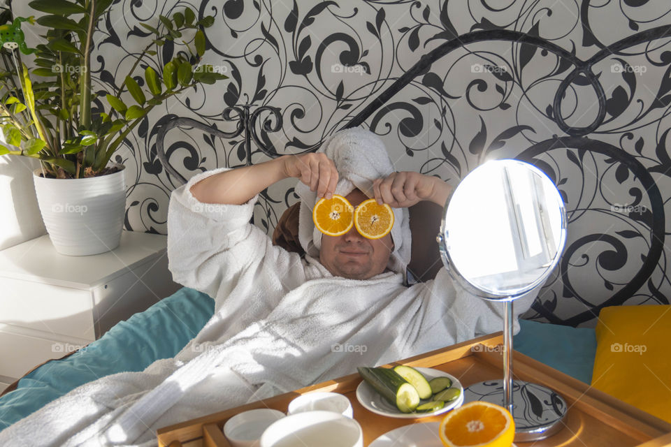 A man and home spa care at home takes care of himself and smears his face with cream, does a manicure at home on a blue background and with oranges and rests.
