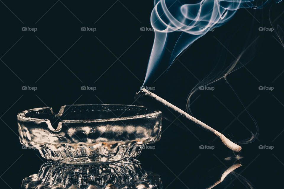 cigar burning in glass ashtray with visible smoke on black background. Studio shot . Low key photo