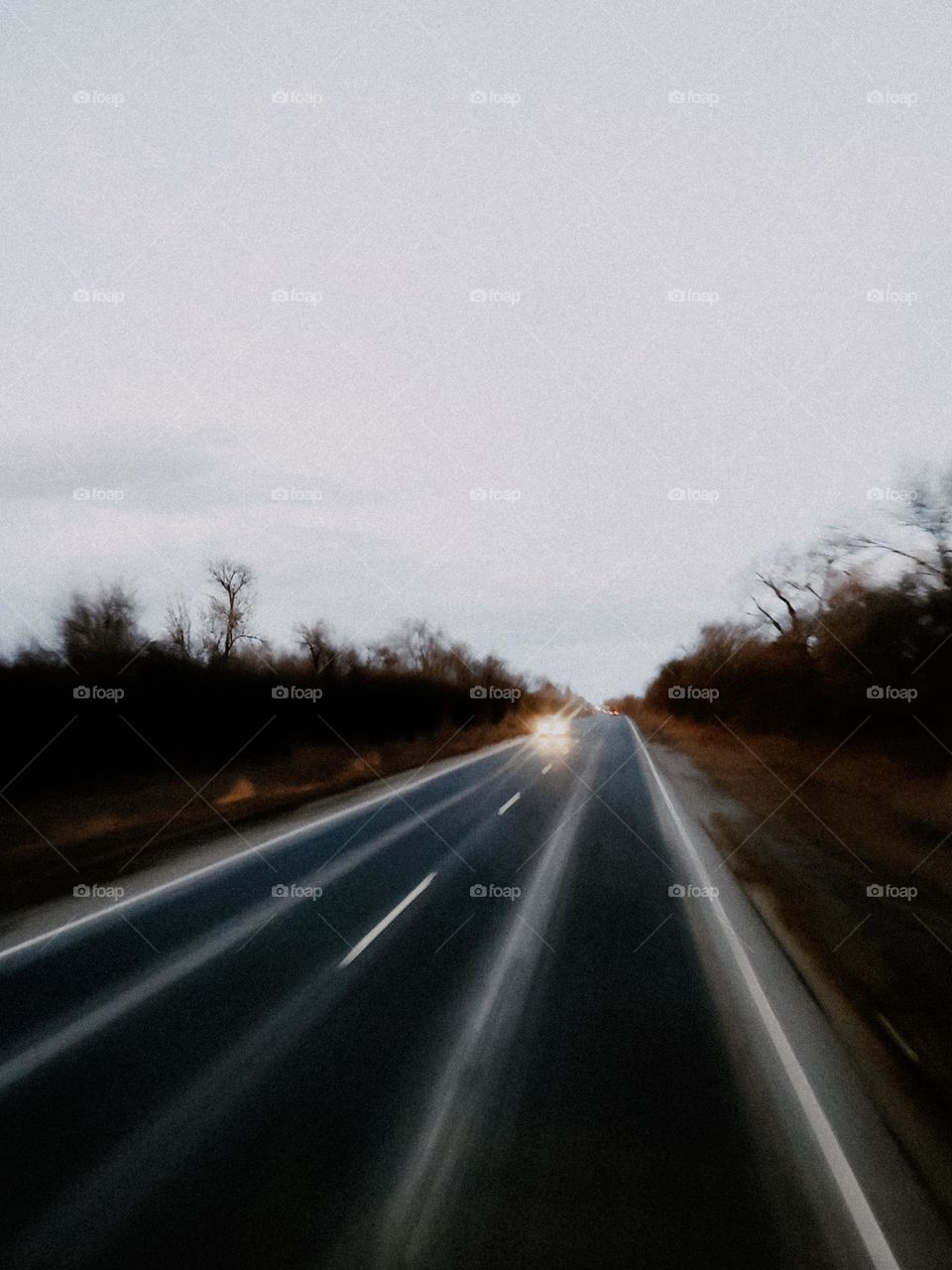 Car with lights on rides on the highway. Car road trip. Gray asphalt highway runs the horizon, trees along the way. Evening sunset sky. Car rides on the road. View from the car window. Eastern Europe, Ukraine
