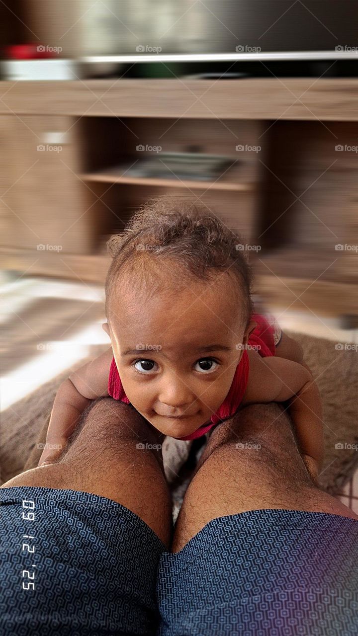 A sweet moment of a baby looking up with big curious eyes, leaning on an adult's legs. The scene captures innocence, affection, and the special bond between child and family in a cozy home setting.