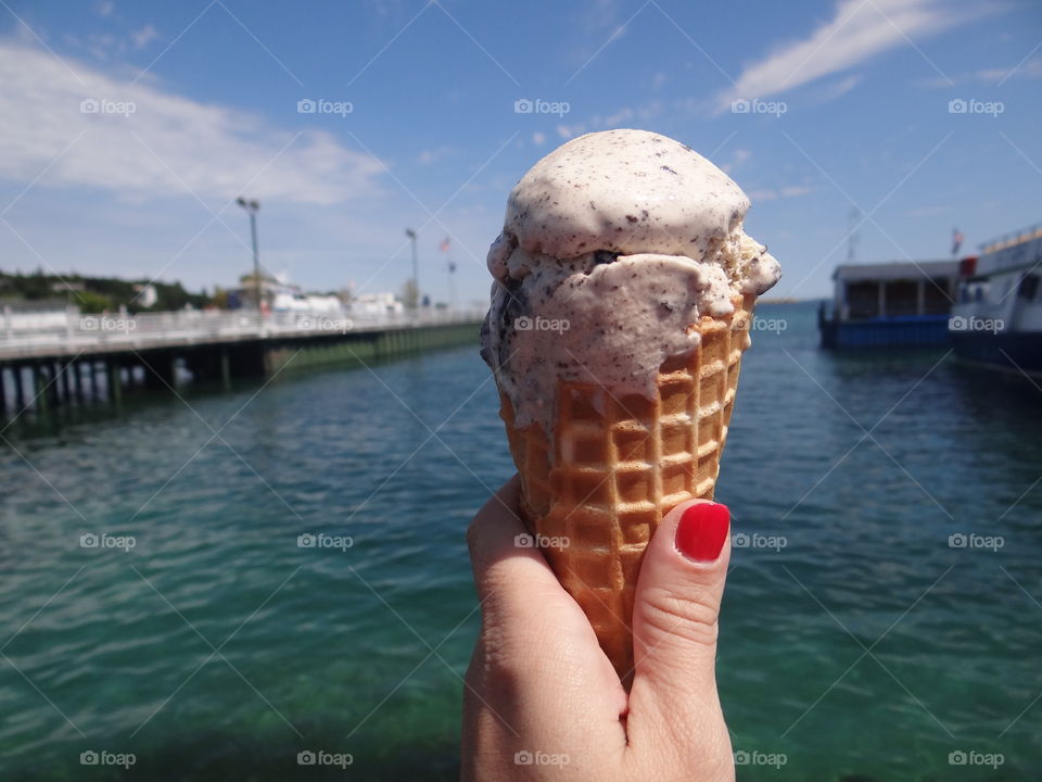 Mackinac Island Ice Cream. Female hand with red nail polish holding waffle cone full of ice cream with Lake Huron in the background.