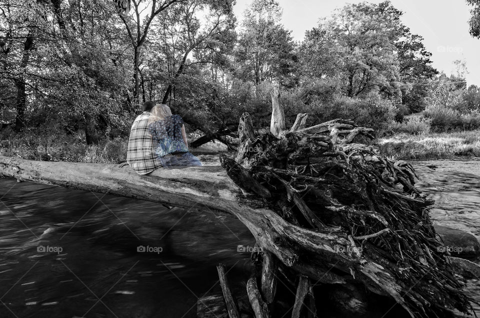 A man sits on the log above a floating river, and the airy silhouette of a woman is seen beside her. The concept of lost loved ones.