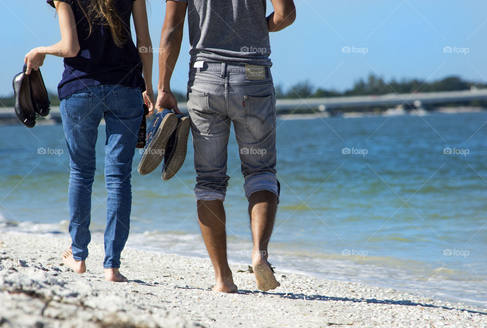 A couple man and woman walk along the beach barefoot with their shoes in their hands by the ocean in summer on vacation