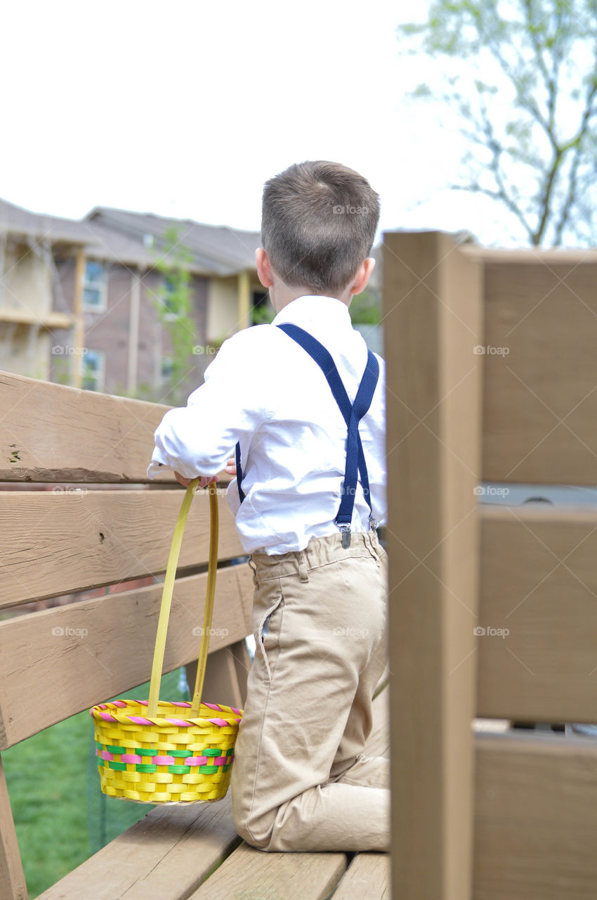 Back view of a young boy wearing suspenders on a bench with an Easter basket looking for Easter eggs