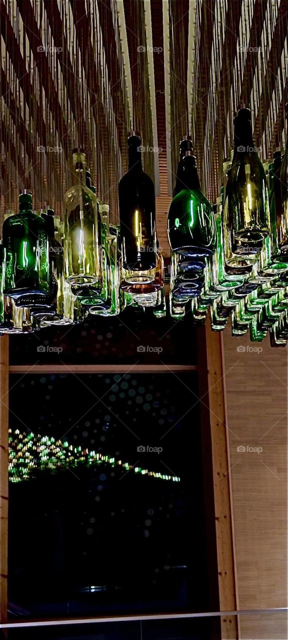 These bottles are suspended from the ceiling with long thin pieces of metal at the „Nawareum“, a concert hall venture in „Straubing“, Germany. They are individually illuminated with electric lights that change over time. 2023. Hypnotic Productions