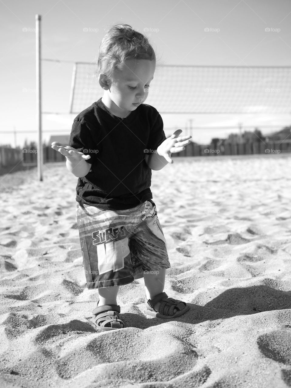 A little toddler boy in shorts and sandals watches curiously as sand falls through his fingers. 