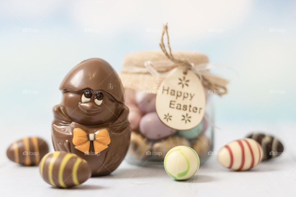 One chocolate chicken in the shell with eggs scattered around the coyette and a glass jar with a sweet treat stand on a white wooden table, close-up side view. Holiday treat concept, happy easter.