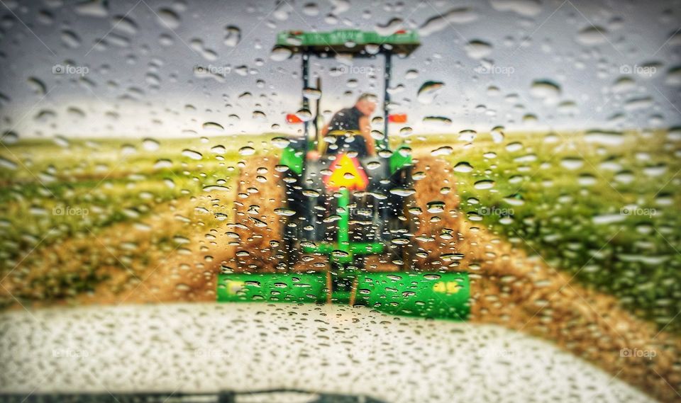 A car being pulled out of the mud by a man driving a John Deere tractor with a chain hooked to it on a country dirt road during a spring rain the photo taken from inside the car thru a windshield with raindrops on it