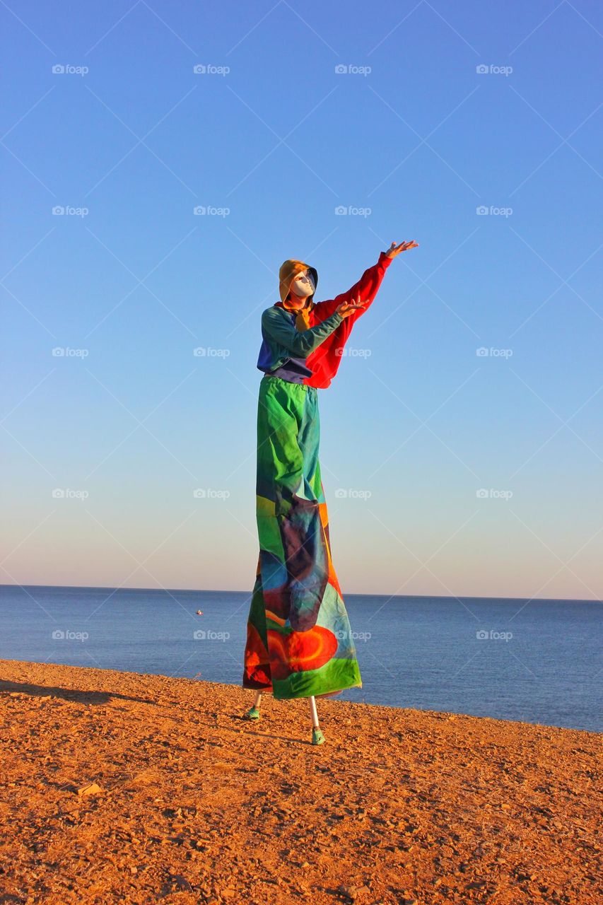 carnival theatrical character on stilts and in a theatrical mask stands on the seashore praising the setting sun