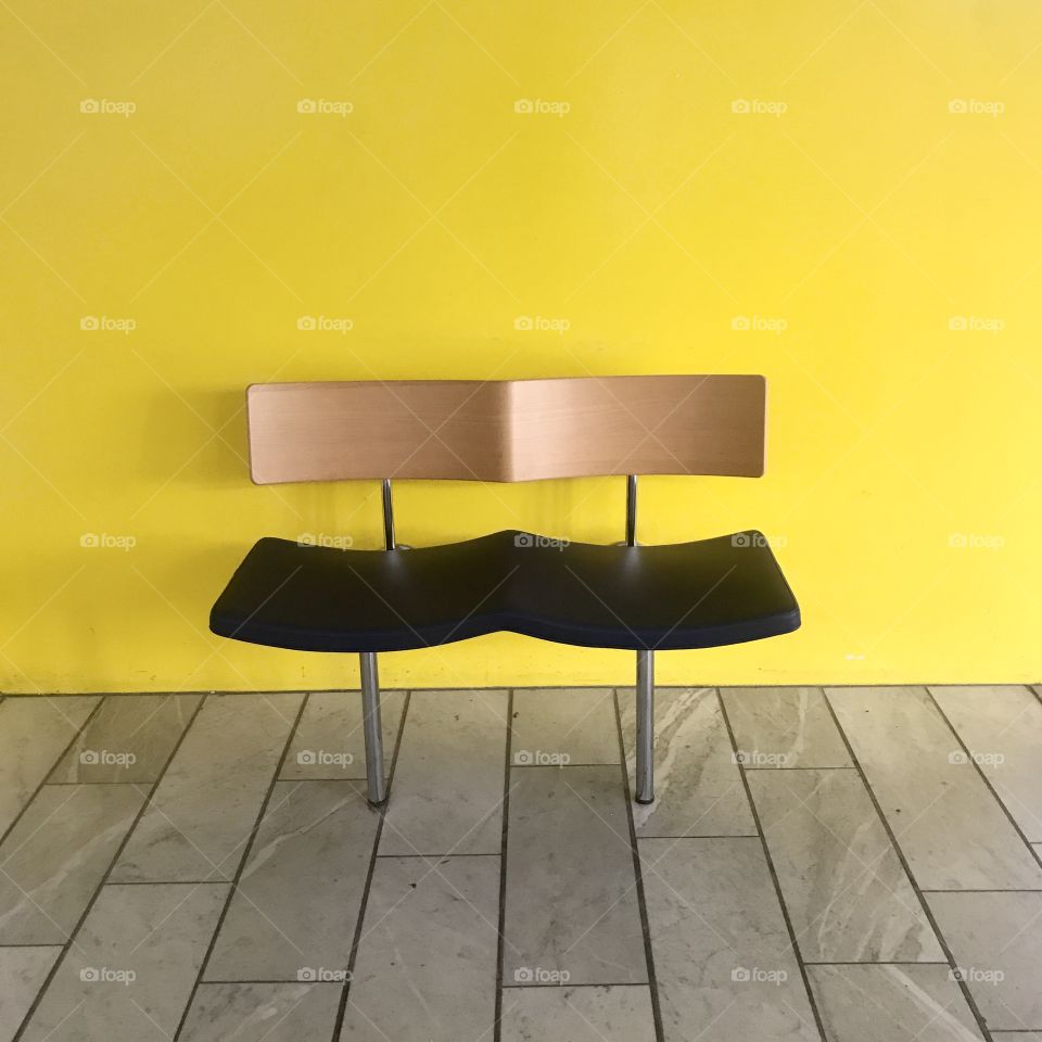Chair bench fitted with two seats set against bright yellow wall and marble floor in gray tones.