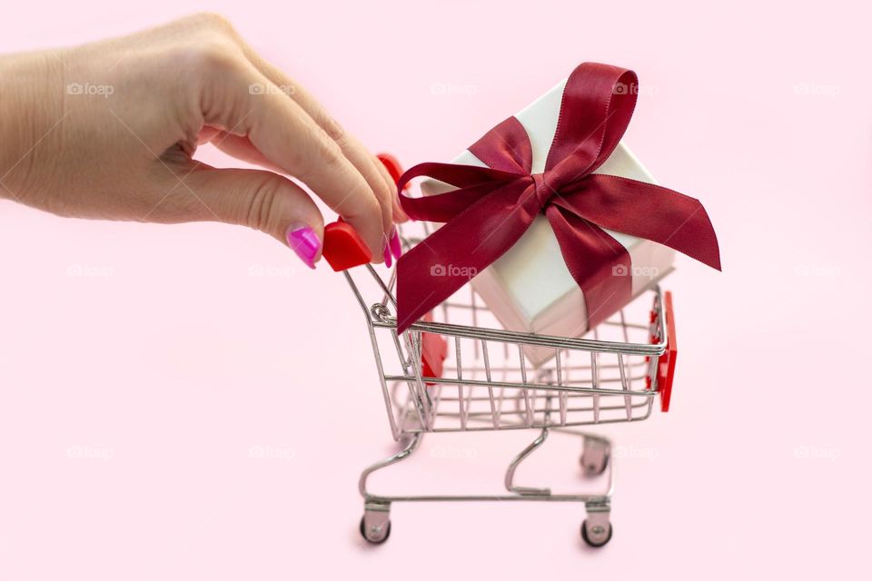 Female hand pushers small shopping cart with the gift box and red bow