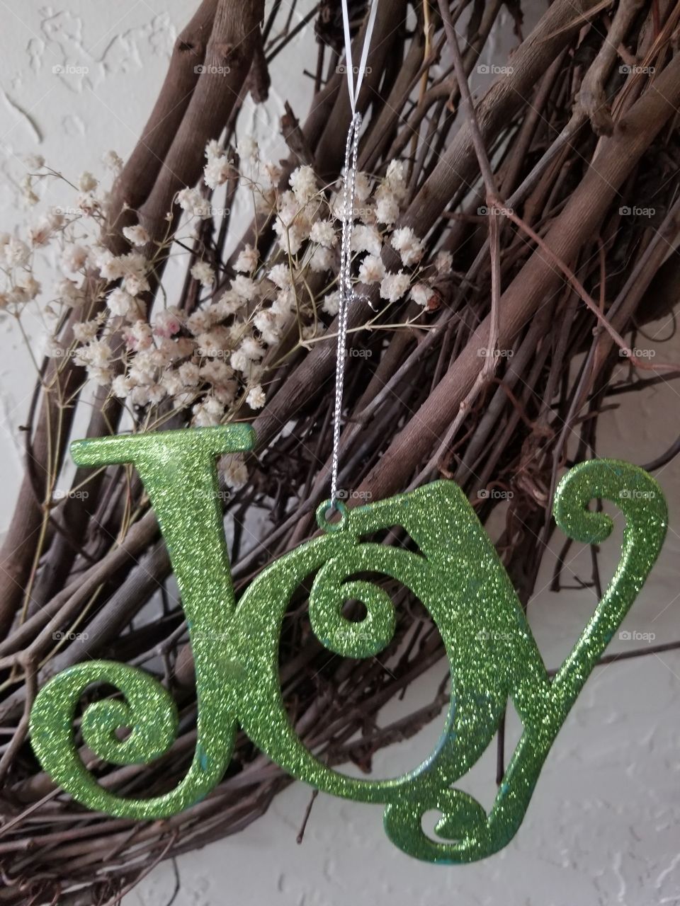 word "joy" in green glitter hangs in front of an intertwined twig wreath adorned with baby breath flowers