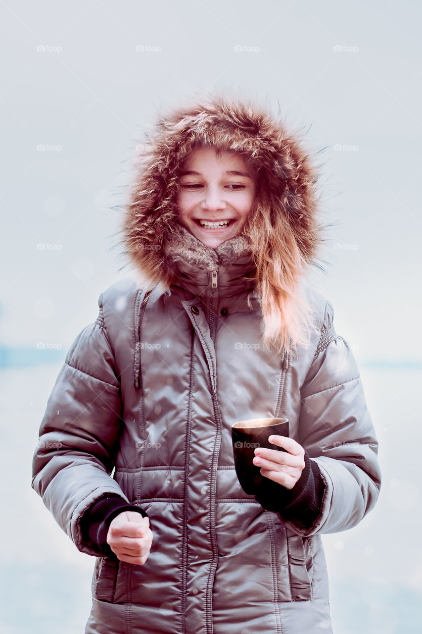 Young pretty smiling girl enjoying drinking hot drink during walk on a wintery day. Wearing coat with hood. Hold a mug of tea