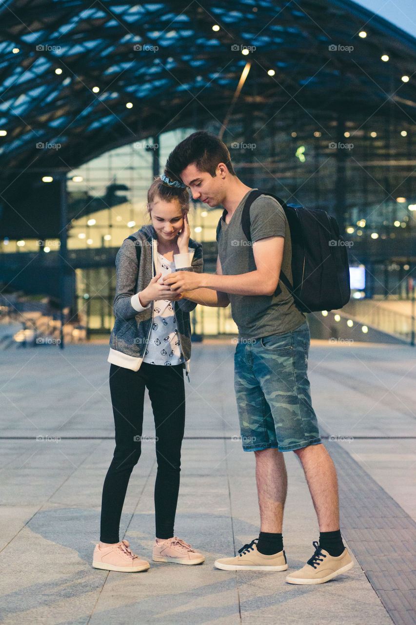 Young woman and man looking at screen of smartphone during walk in the city at night