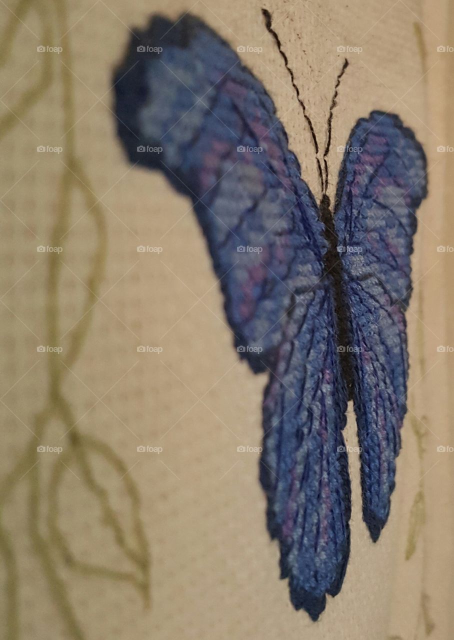 Close up of a blue-hued cross-stitched butterfly, side angled view.