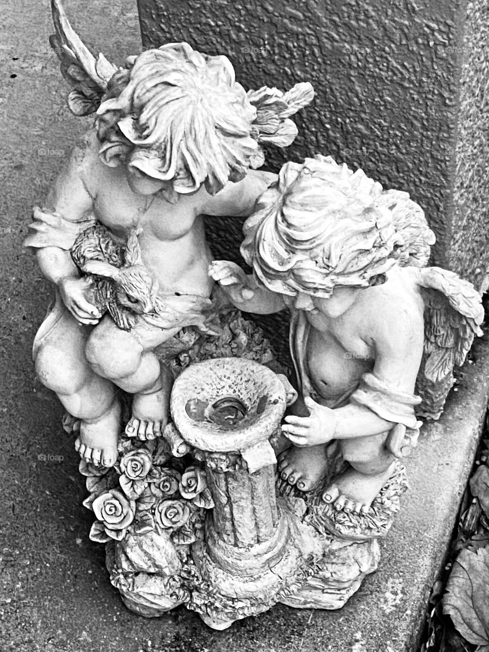 A small resin statue of 2 cherubs with a bird bath and roses and a rabbit