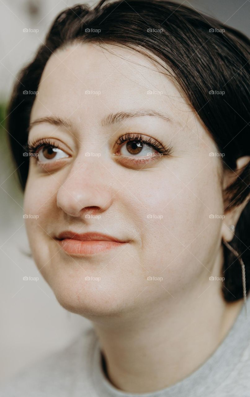 Portrait of one beautiful Caucasian brunette girl with minimal makeup smiling looking away sitting indoors on a summer day, close-up view from the bottom up on the side.