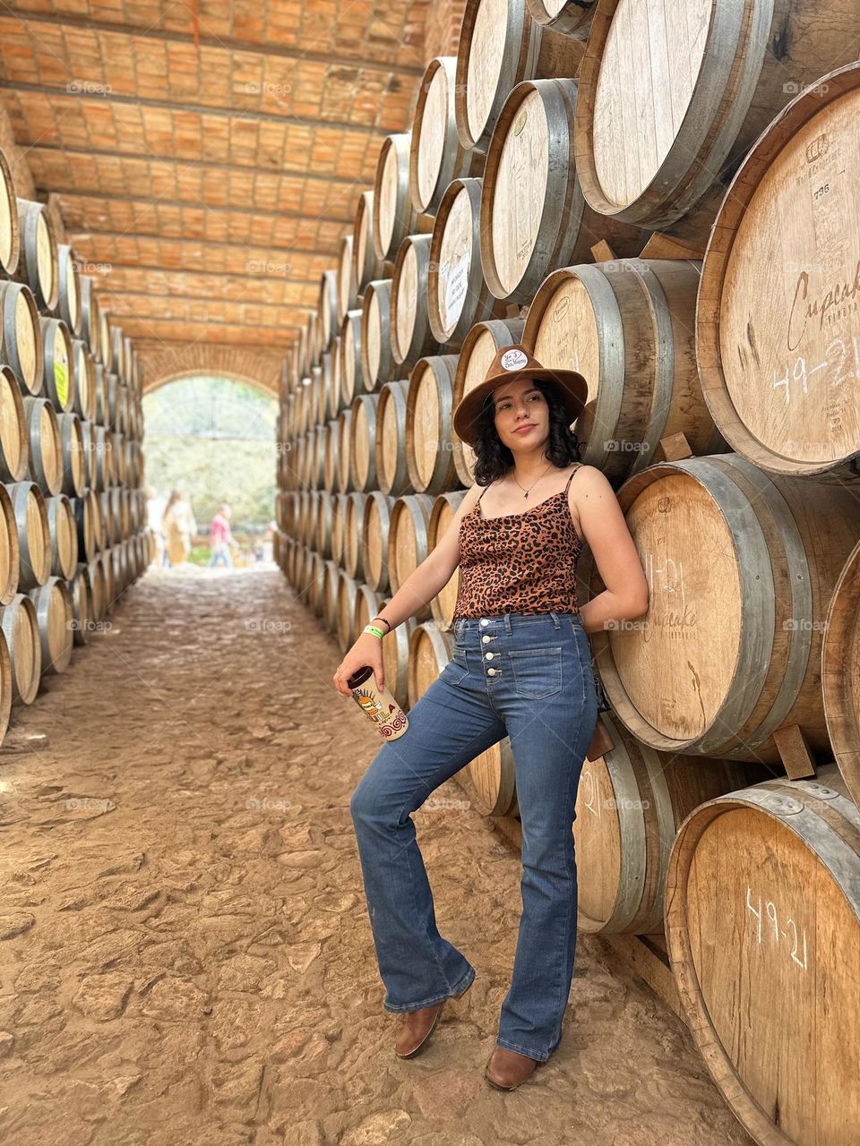 Woman wearing a hat, leaning on barrels while holding a drink with her hand.