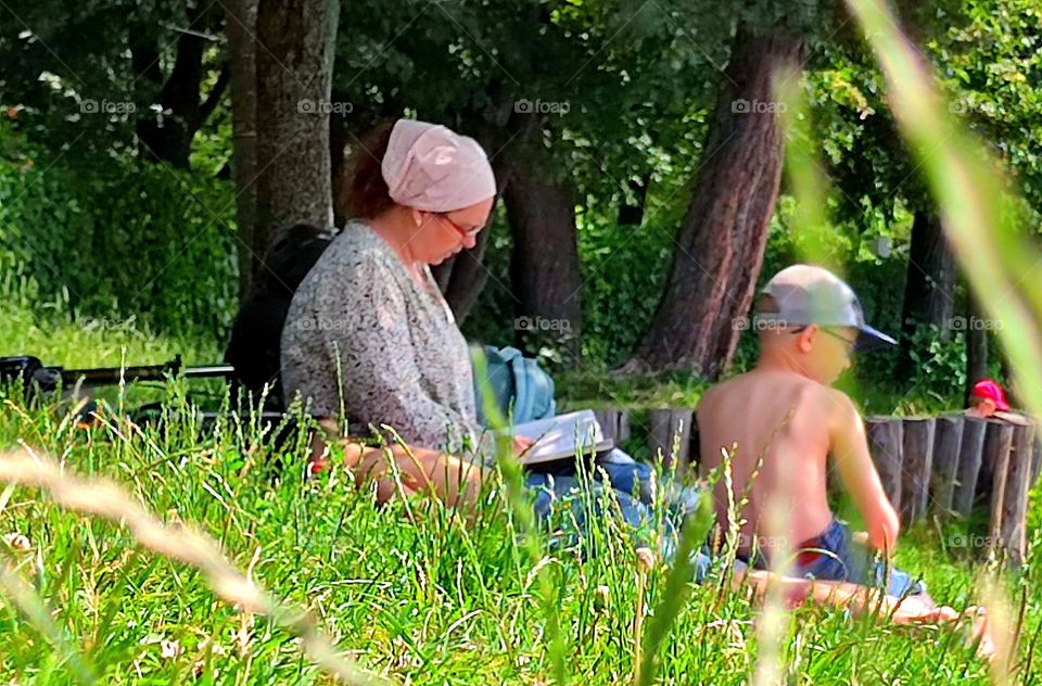 Summer in the city.  A park.  A woman sits on the green grass under the shade of a tree and reads a book to two boys